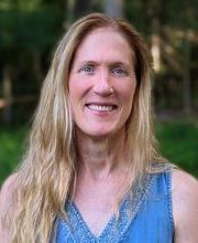 Dr. Darcy Boellstorf smiling with long straight dirty blond hair wearing a v-neck sleeveless blue top with grass and trees in photo background