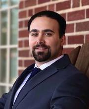 Dr. Kianoosh Hassani smiling with short dark brown hair, mustache and beard and wearing a navy blue suit jacket and tie with button down white shirt